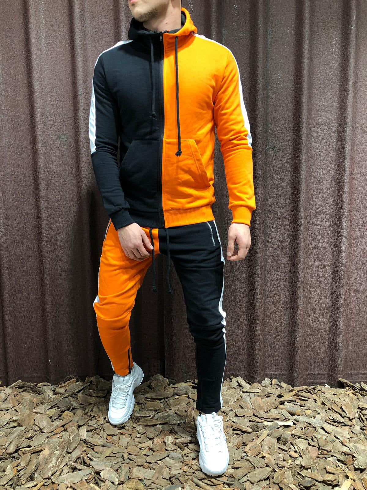 Uomo con tuta sportiva bicolore arancione e nera, cappuccio e pantaloni abbinati, scarpe bianche, sfondo marrone. Moda casual maschile.