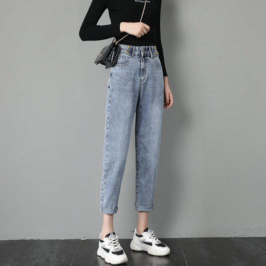 Donna in jeans a vita alta e maglia nera, con borsa a tracolla e sneakers bianche, posa contro un muro grigio. Moda casual femminile.