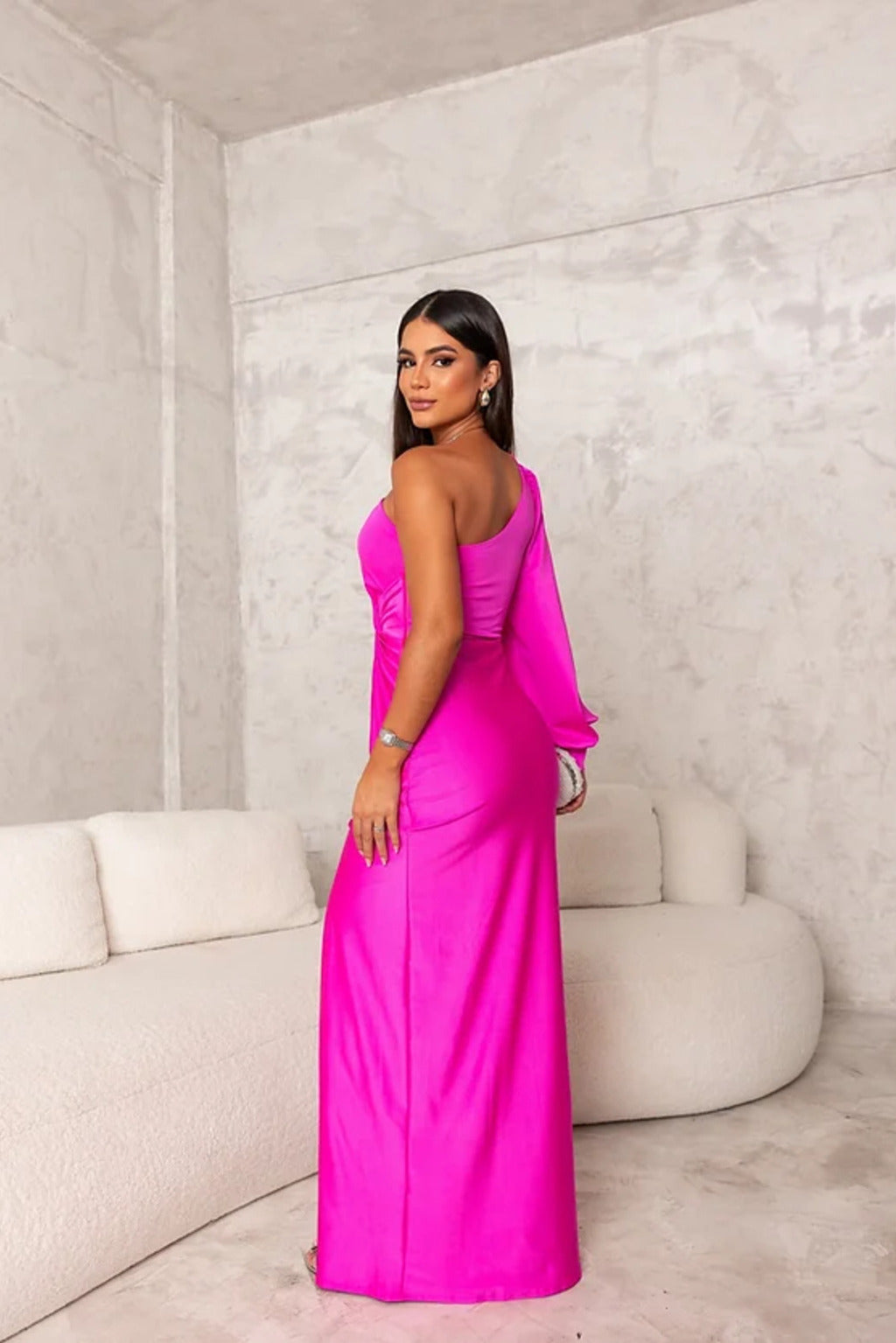 Donna in abito lungo rosa acceso, elegante e moderno, posa in un salotto minimalista con divano bianco. Moda femminile, stile glamour.