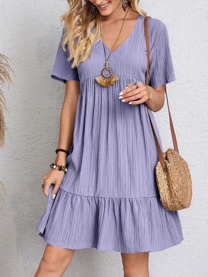 Donna con abito estivo viola, tessuto a pieghe, borsa a tracolla in paglia, accessori boho. Moda casual femminile, look estivo elegante.