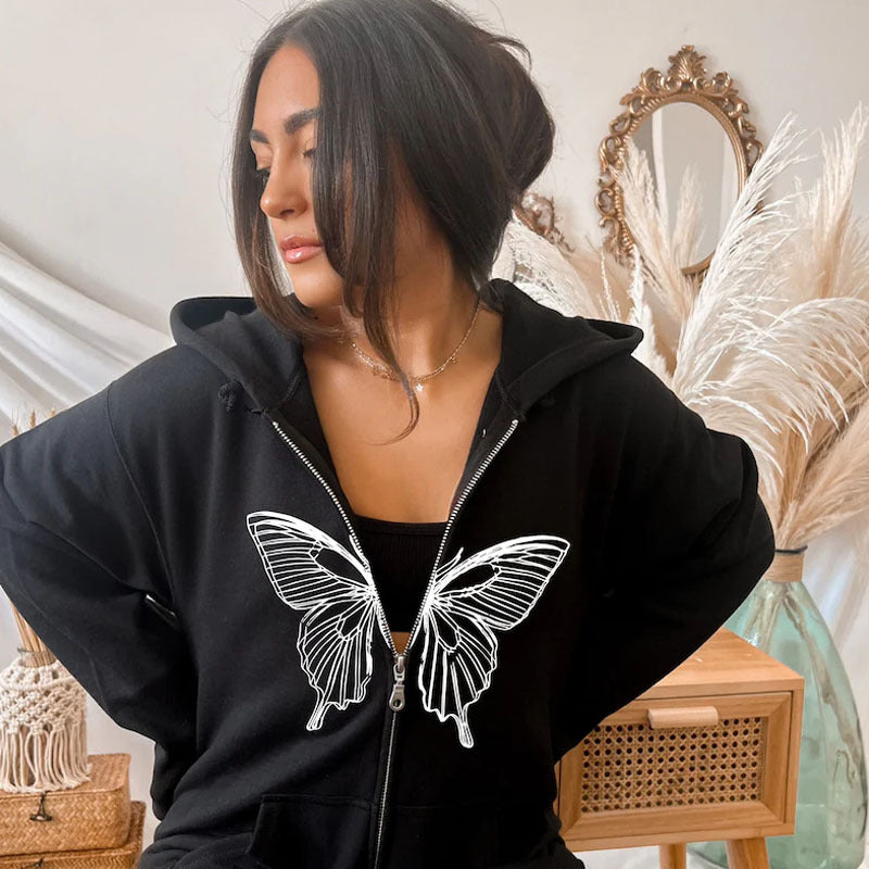 Donna con felpa nera con farfalla bianca, capelli scuri raccolti, sfondo con piante di pampas e specchio decorativo. Moda casual autunnale.