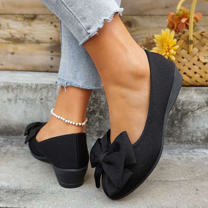 Scarpe ballerine nere con fiocco, indossate con jeans chiari strappati alla caviglia, accanto a un cesto di fiori gialli.