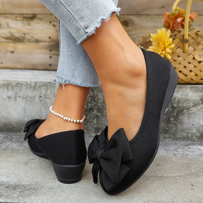 Scarpe ballerine nere con fiocco, indossate con jeans chiari strappati alla caviglia, accanto a un cesto di fiori gialli.