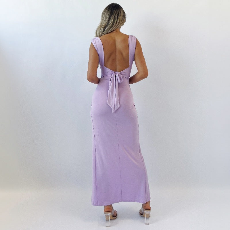 Donna in abito lungo lilla con schiena scoperta e fiocco, sandali trasparenti. Elegante vestito da sera, moda femminile, tendenze abbigliamento 2023.