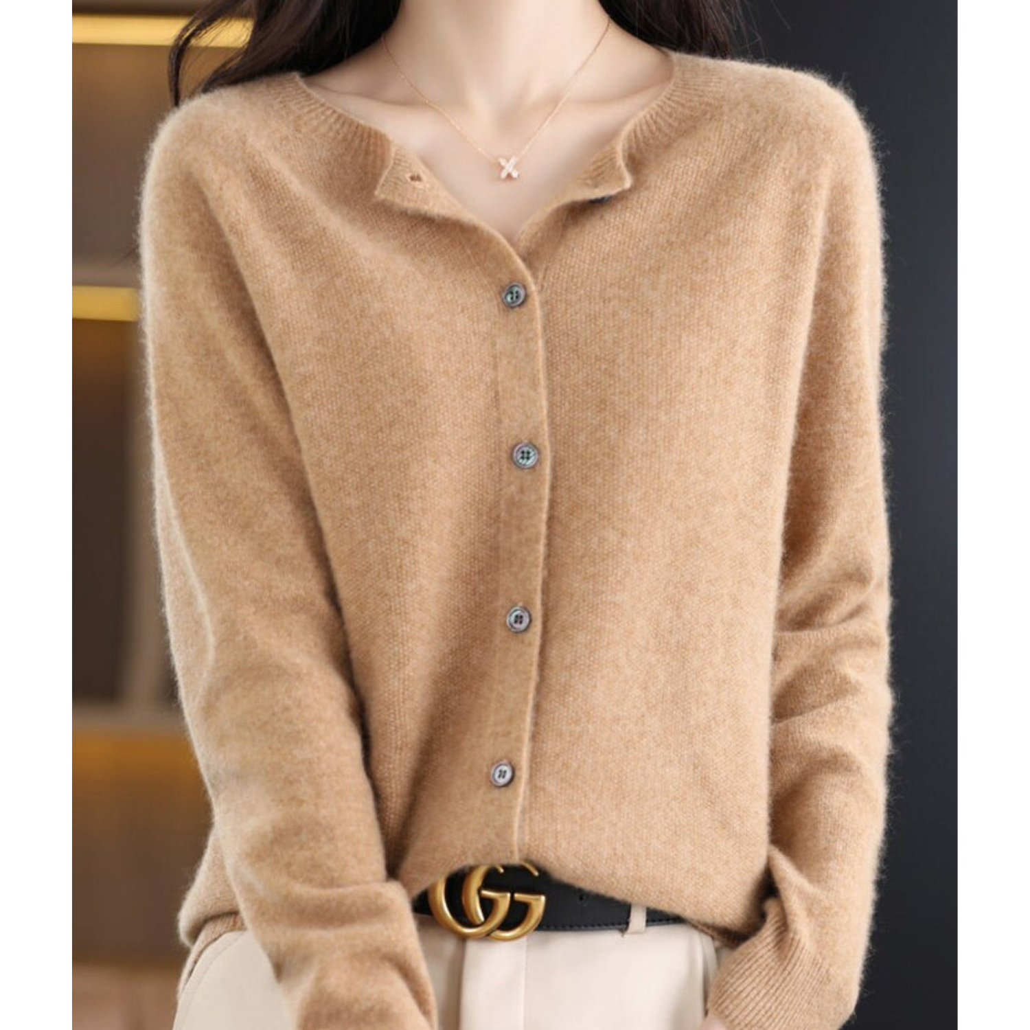 Maglione beige in cashmere con bottoni, indossato da una donna. Accessori: collana con ciondolo a X e cintura nera con fibbia dorata.