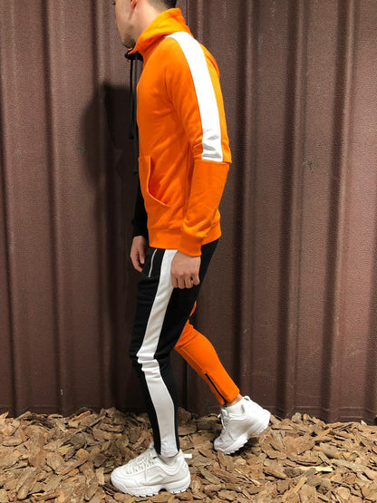 Uomo in tuta sportiva arancione e nera con strisce bianche, scarpe da ginnastica bianche, cammina su terreno di corteccia. Moda casual maschile.