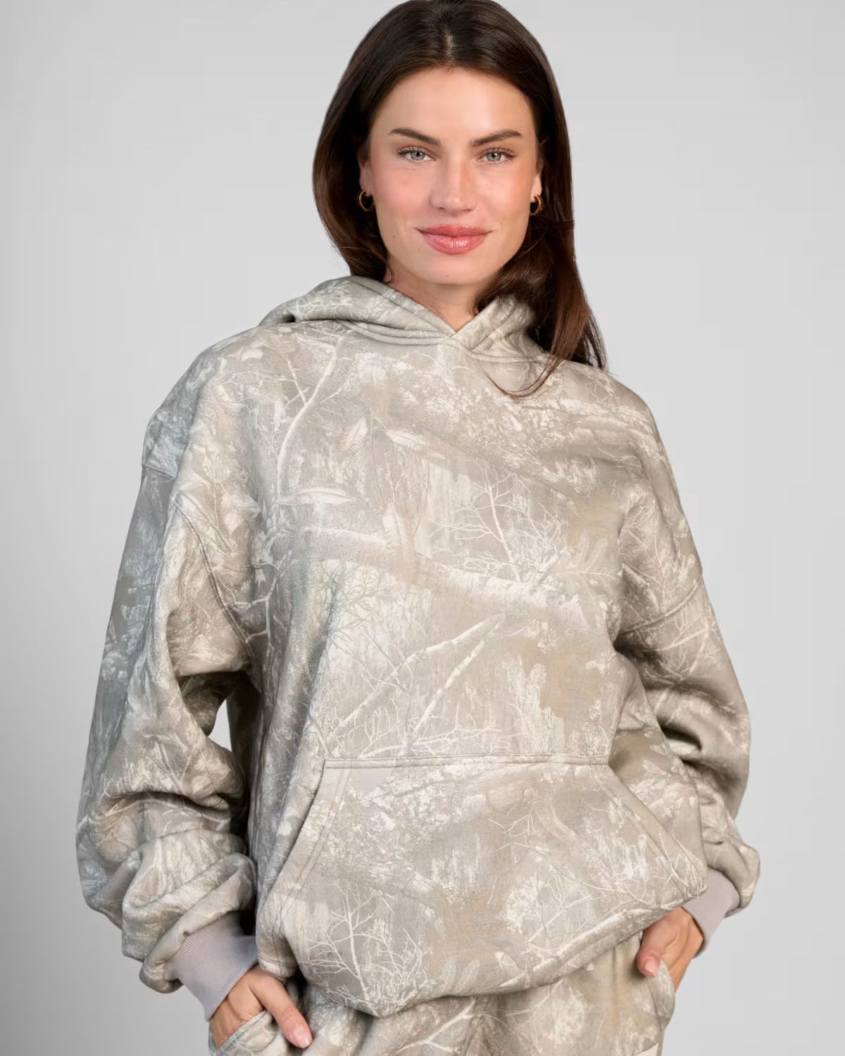 Donna con felpa oversize mimetica beige, mani in tasca, sfondo grigio. Moda casual, abbigliamento autunnale, stile urbano.