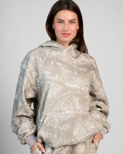 Donna con felpa oversize mimetica beige, mani in tasca, sfondo grigio. Moda casual, abbigliamento autunnale, stile urbano.