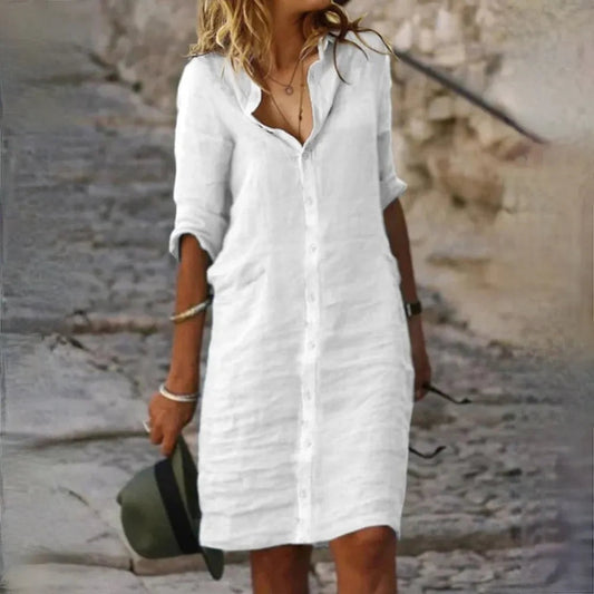 Donna con abito bianco di lino, maniche lunghe, cammina su strada acciottolata. Moda estiva casual, stile elegante, accessori minimal.