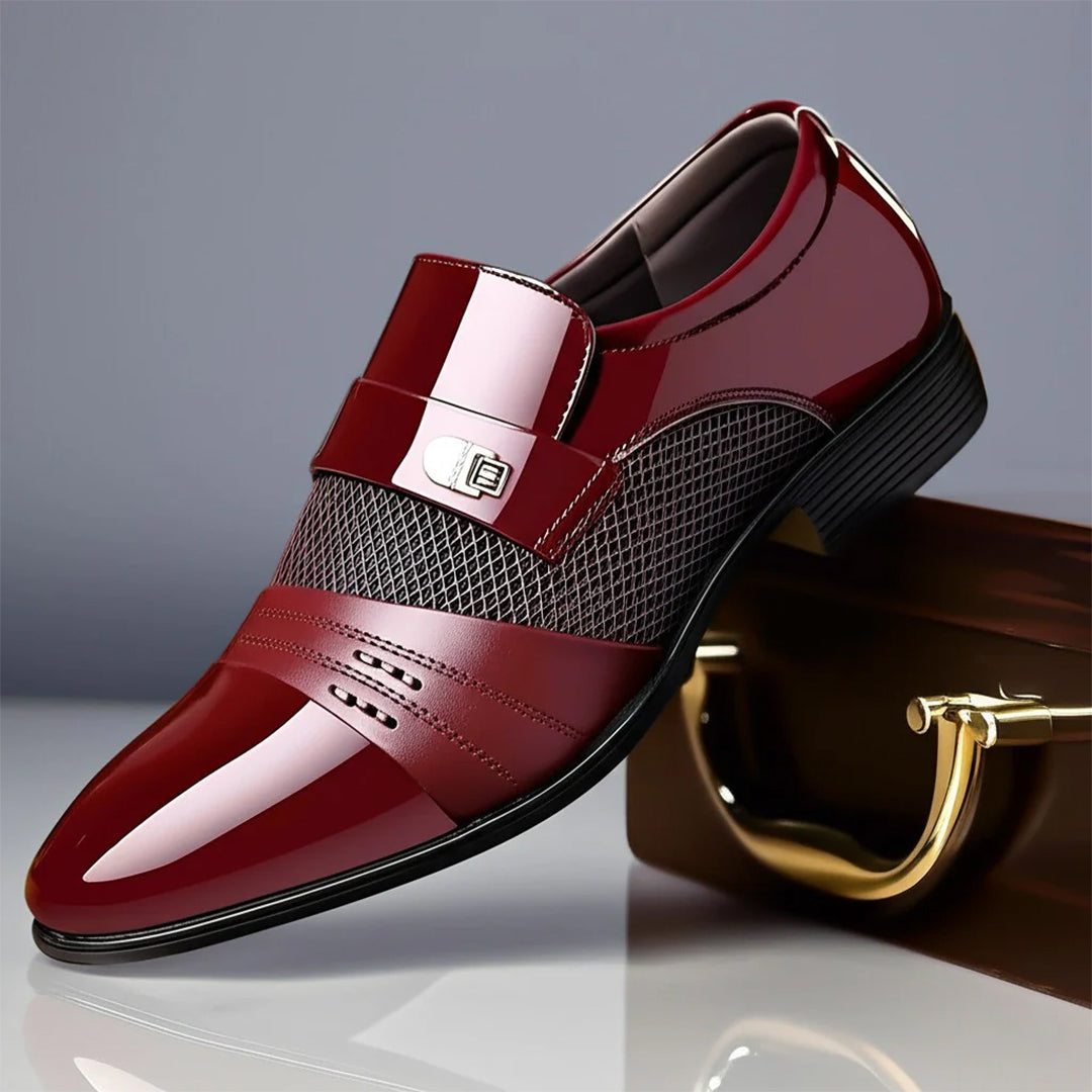 Scarpa elegante da uomo in pelle lucida bordeaux con fibbia metallica, design traforato e suola nera, posata su una valigetta marrone.