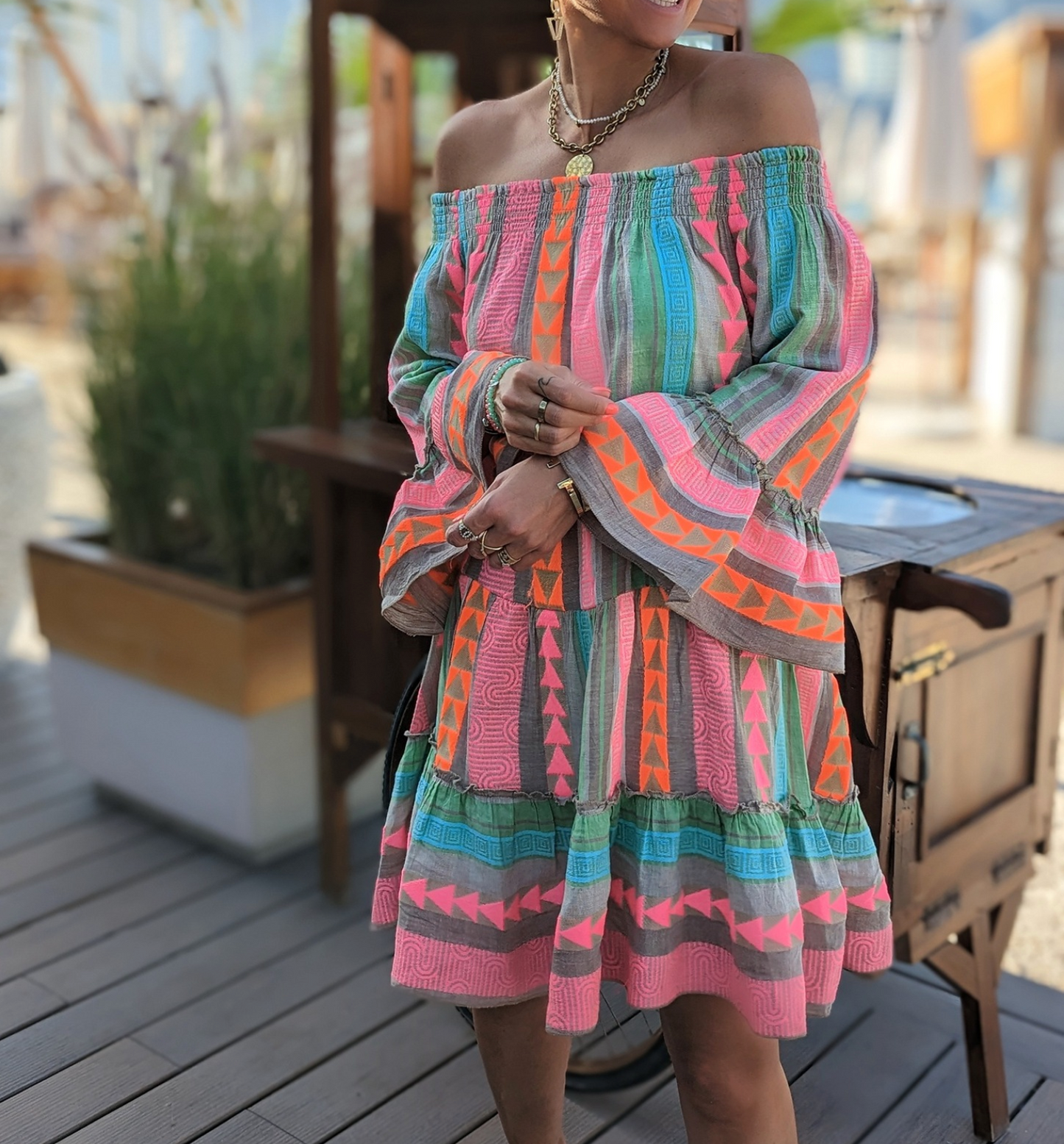 Abito estivo colorato con motivi geometrici, maniche lunghe e spalle scoperte, indossato da una donna su una terrazza all'aperto.