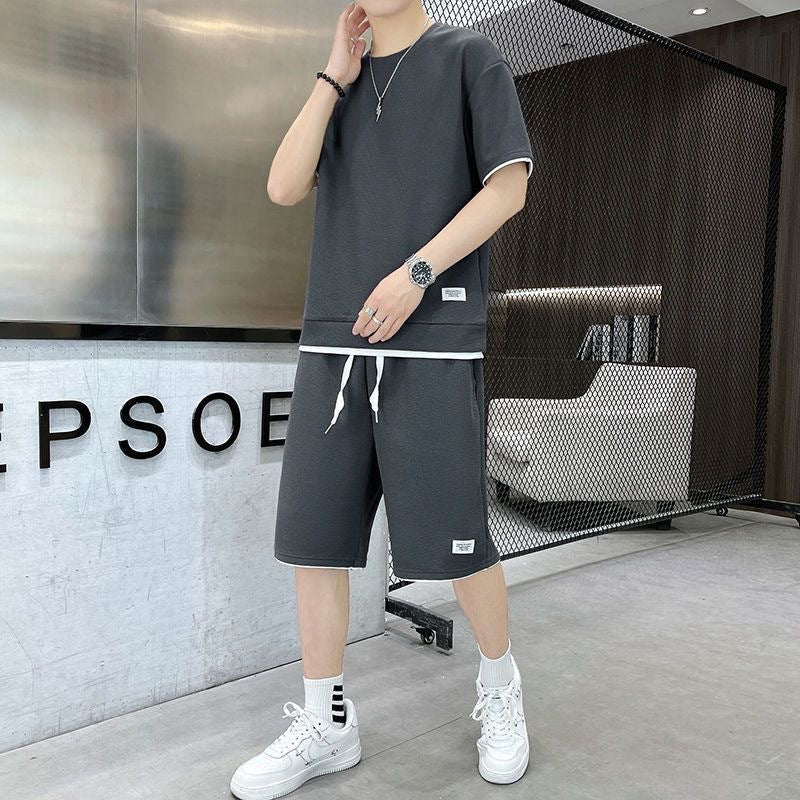 Giovane uomo in abbigliamento casual grigio, t-shirt e pantaloncini, con scarpe da ginnastica bianche, in un ambiente moderno e minimalista.