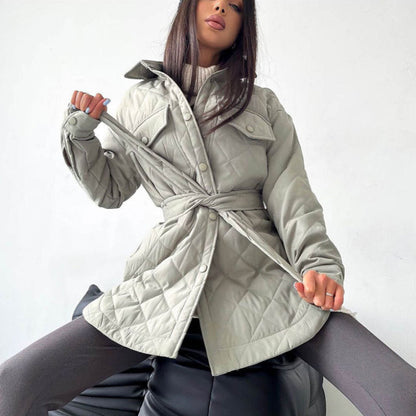 Donna con cappotto trapuntato grigio chiaro, cintura in vita, seduta su poltrona nera. Moda autunnale, stile elegante, abbigliamento femminile.