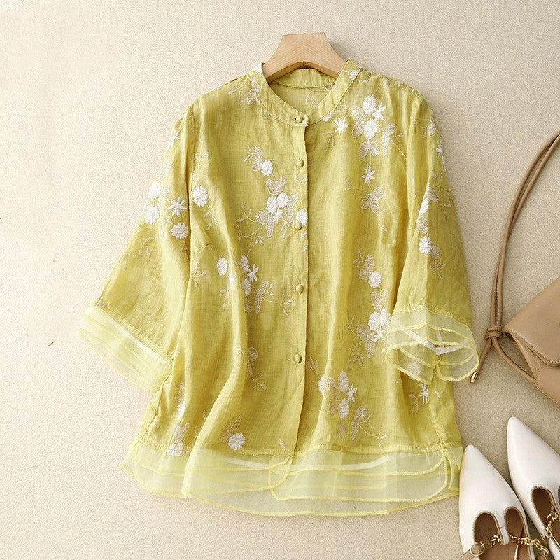 Camicia gialla in lino con ricami floreali bianchi, maniche a tre quarti, su sfondo beige. Moda estiva donna, abbigliamento casual elegante.