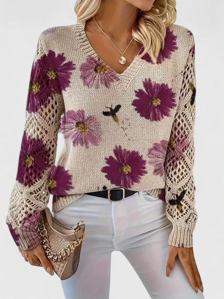 Maglione donna beige con fiori viola, maniche traforate, abbinato a pantaloni bianchi e borsa a catena. Stile casual chic autunnale.