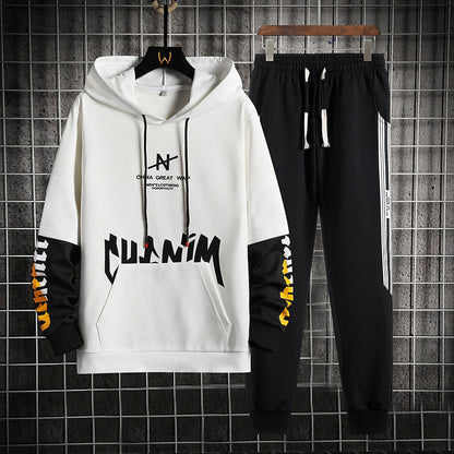 Set tuta sportiva uomo, felpa bianca con cappuccio e scritte nere, pantaloni neri con strisce laterali bianche, appeso su griglia metallica.