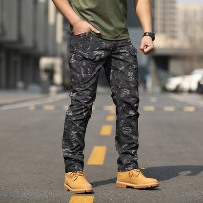 Uomo in piedi su strada indossando pantaloni mimetici neri e scarpe gialle, sfondo urbano sfocato. Moda maschile casual, stile urbano.