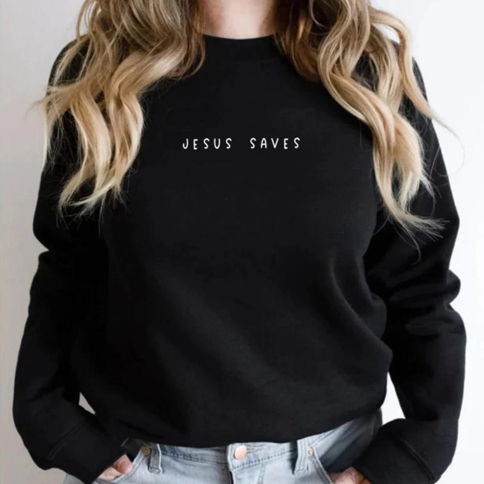 Donna con maglione nero "Jesus Saves", capelli biondi ondulati, jeans chiari. Abbigliamento casual, moda cristiana, messaggio religioso.