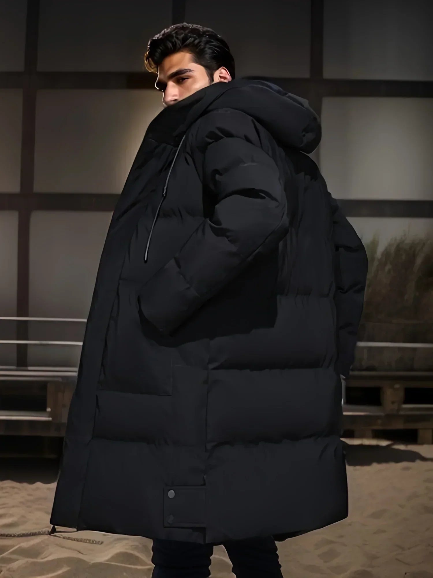 Giovane uomo con cappotto invernale nero imbottito, cappuccio, in piedi su una spiaggia di sera. Moda maschile, abbigliamento invernale elegante.