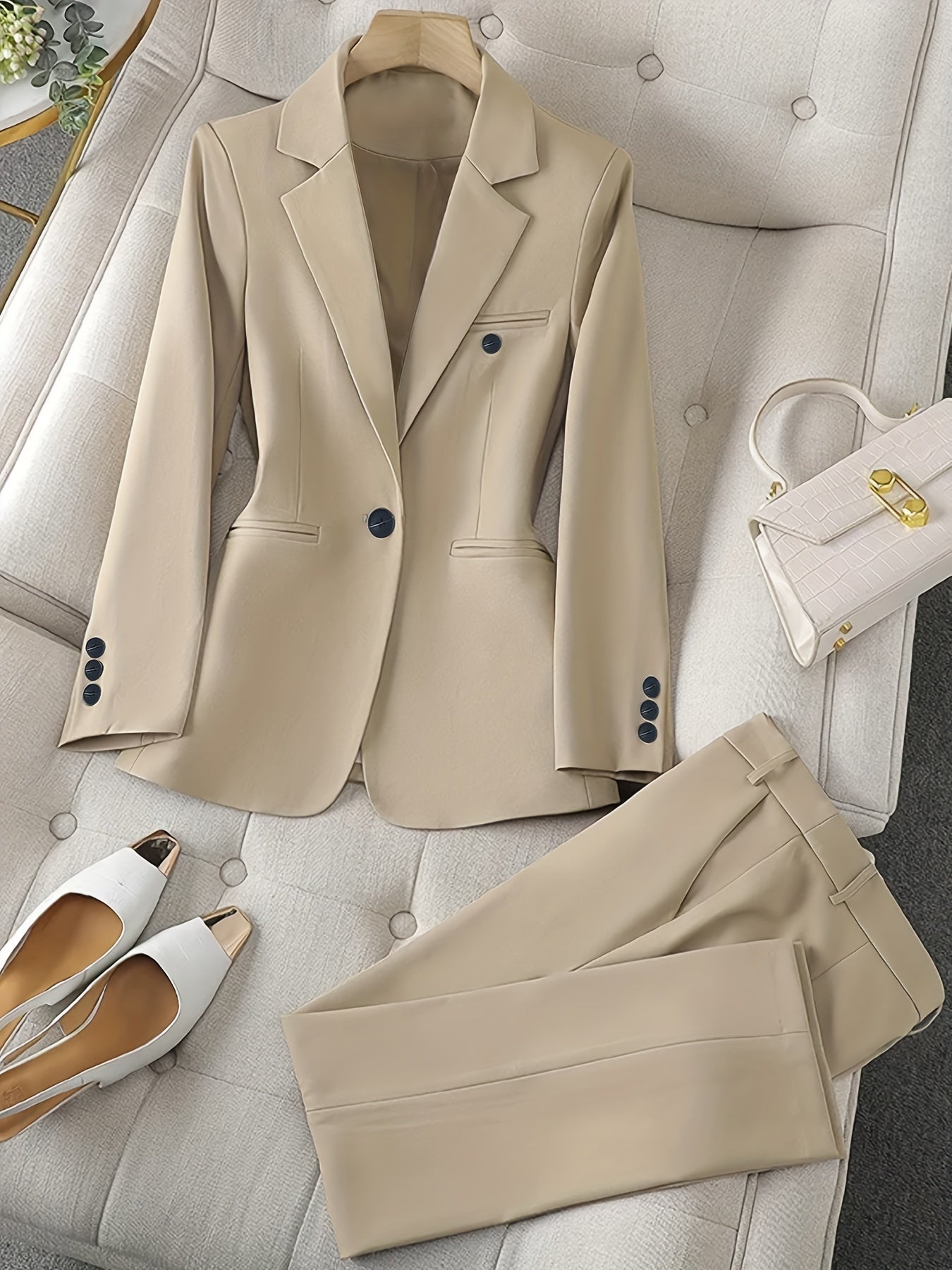 Completo elegante beige da donna su divano, con giacca e pantaloni abbinati, borsa bianca e scarpe col tacco. Moda femminile, abbigliamento formale.