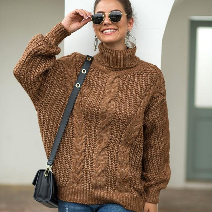 Donna sorridente con occhiali da sole indossa maglione marrone a trecce, borsa a tracolla nera, orecchini grandi, posa all'aperto. Moda autunnale casual.