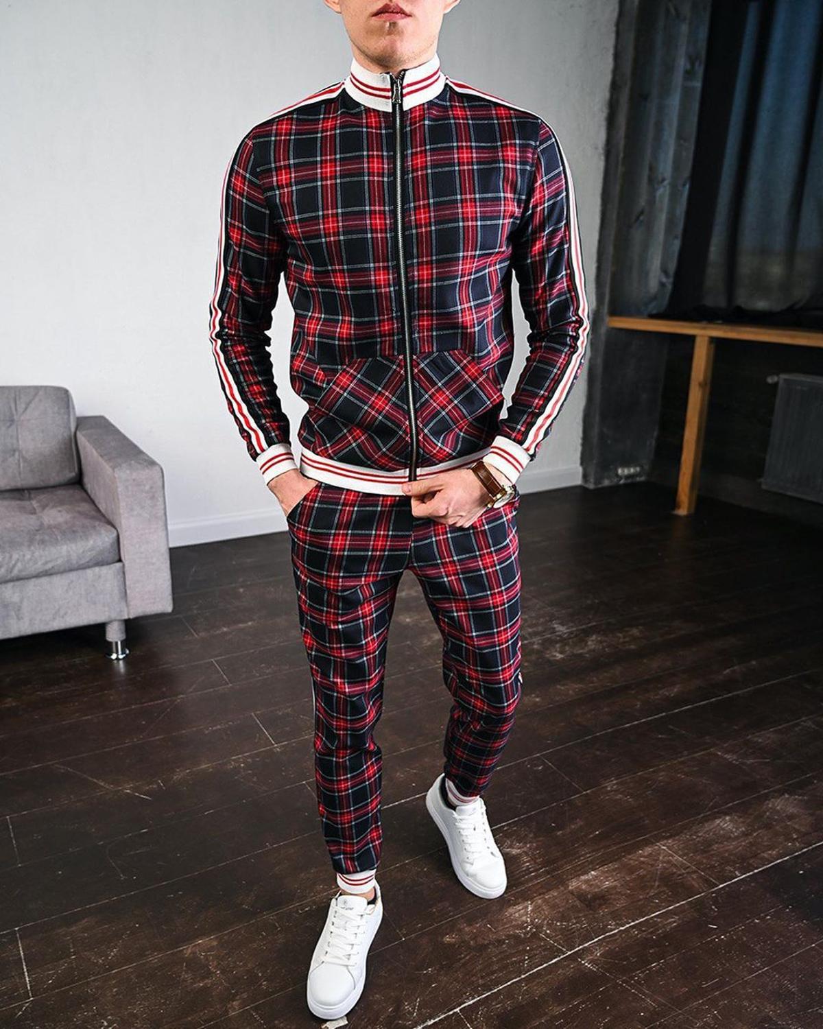 Tuta sportiva a quadri rossi e neri, uomo in piedi su pavimento in legno, scarpe bianche, divano grigio sullo sfondo. Moda casual maschile.
