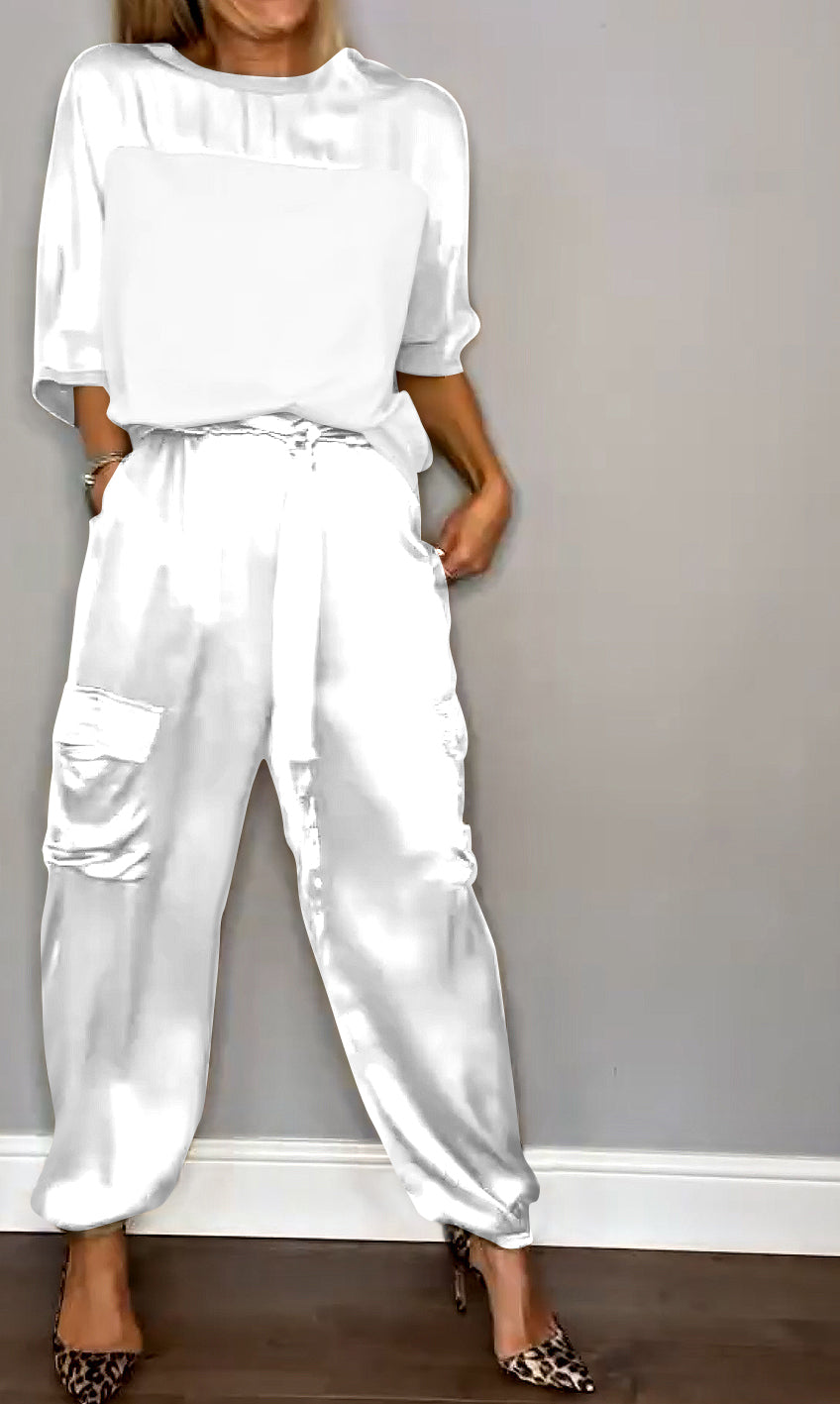 Donna in abbigliamento elegante bianco, indossa pantaloni larghi con tasche e camicetta, scarpe leopardate, posa contro parete grigia.