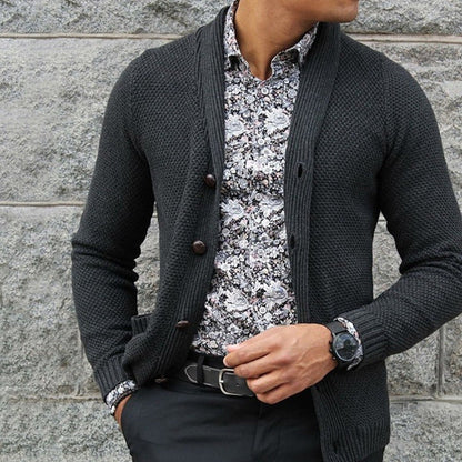 Uomo con cardigan grigio scuro, camicia a fiori bianca e nera, pantaloni neri. Moda maschile elegante, stile autunnale, outfit casual chic.
