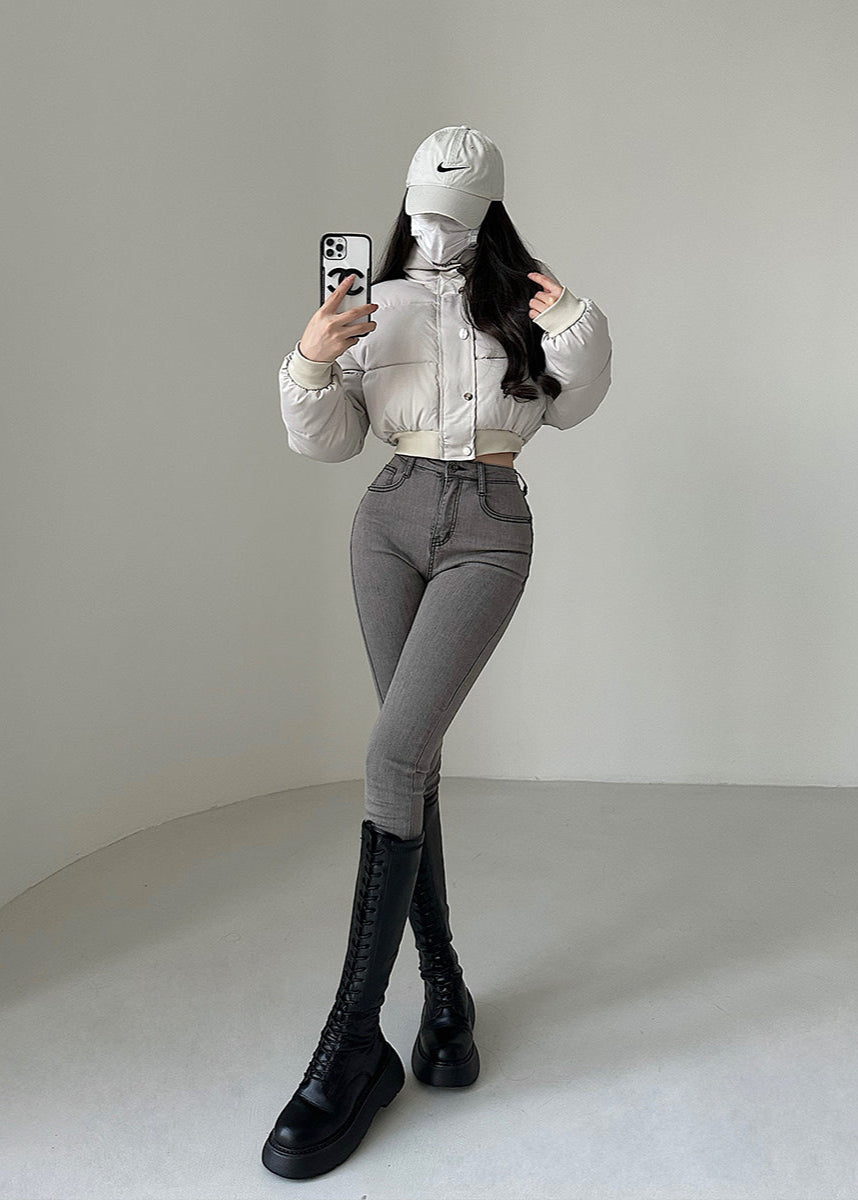 Donna con cappello e mascherina, indossa giacca bianca, jeans grigi e stivali neri, scatta selfie con smartphone in una stanza minimalista.