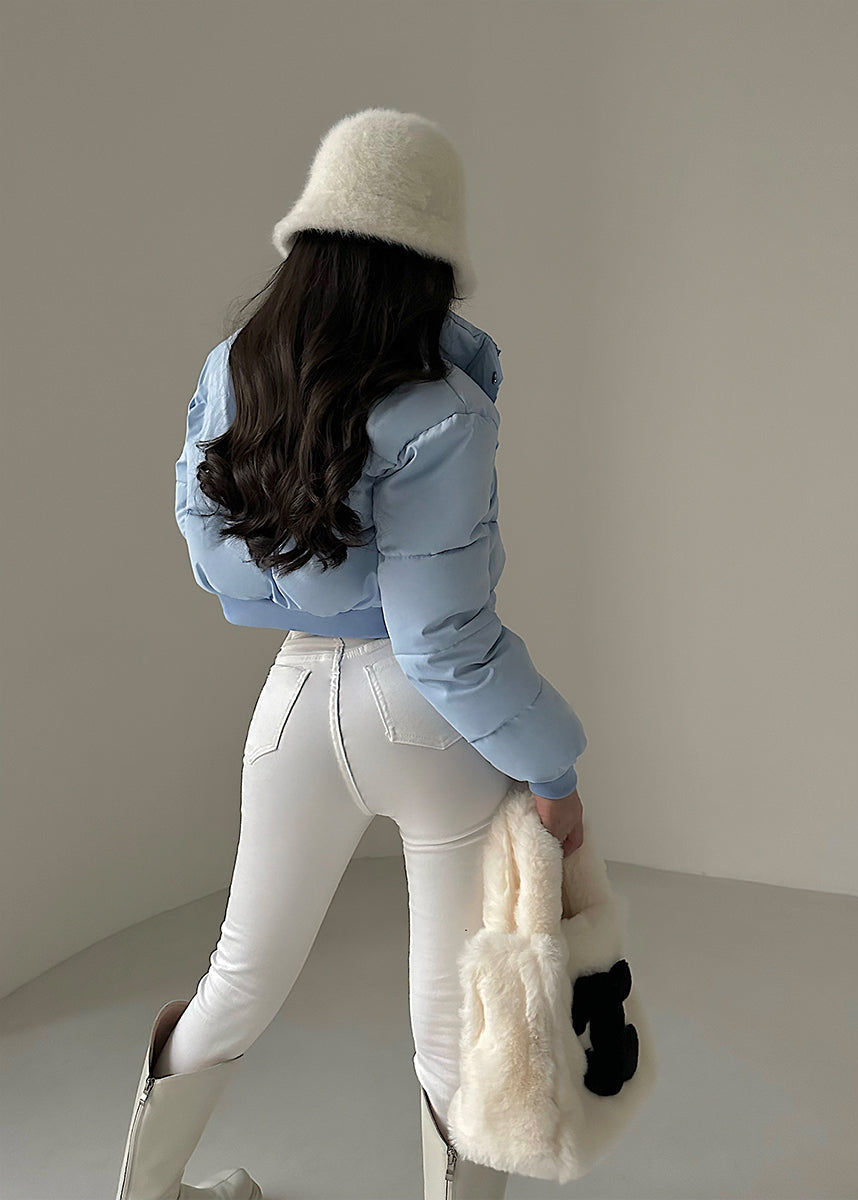 Donna di spalle con cappotto azzurro, pantaloni bianchi, stivali alti e cappello di pelliccia bianca. Borsa di pelliccia bianca in mano.