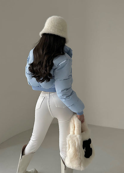 Donna di spalle con cappotto azzurro, pantaloni bianchi, stivali alti e cappello di pelliccia bianca. Borsa di pelliccia bianca in mano.