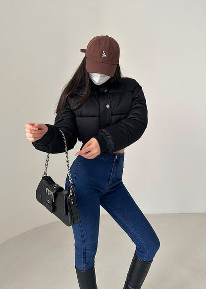 Donna con giacca nera imbottita, jeans attillati blu, stivali neri, cappello marrone e mascherina bianca, tiene una borsa a tracolla nera.