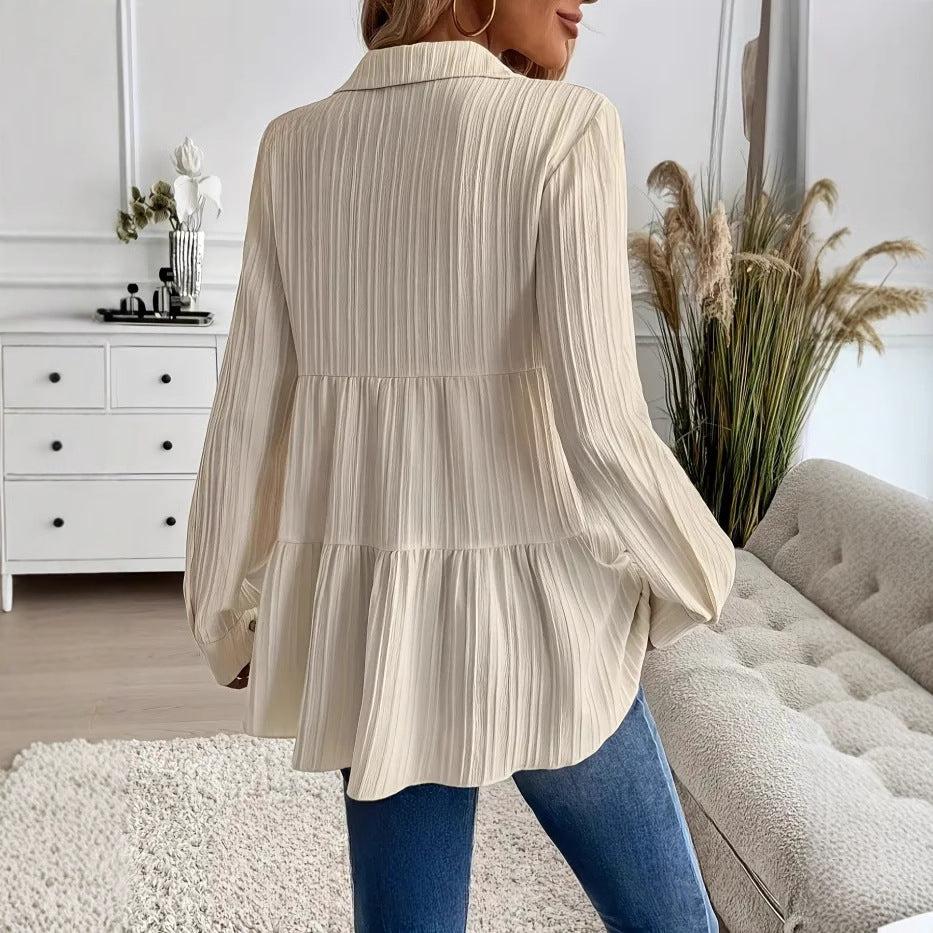 Donna con camicia plissettata beige, jeans blu, in salotto moderno con divano grigio e piante decorative. Moda casual elegante, arredamento minimalista.