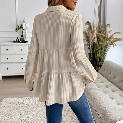 Donna con camicia plissettata beige, jeans blu, in salotto moderno con divano grigio e piante decorative. Moda casual elegante, arredamento minimalista.