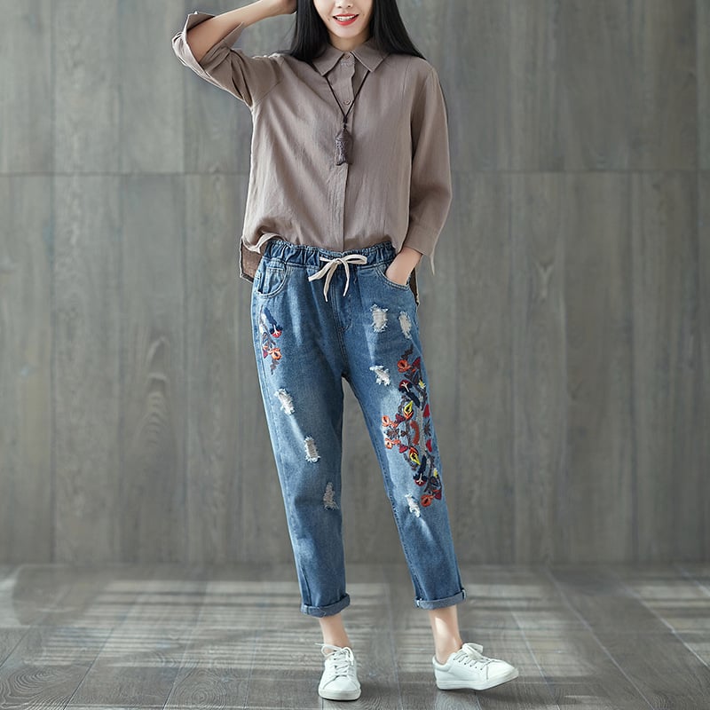 Donna in camicia marrone e jeans ricamati, posa su sfondo in legno. Moda casual, abbigliamento femminile, stile urbano, scarpe bianche.