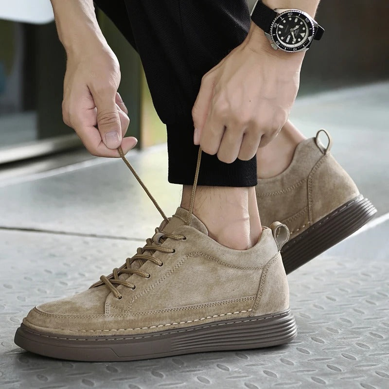 Scarpe da uomo in pelle scamosciata beige, design casual, suola marrone, lacci, indossate con pantaloni neri. Orologio da polso nero visibile.