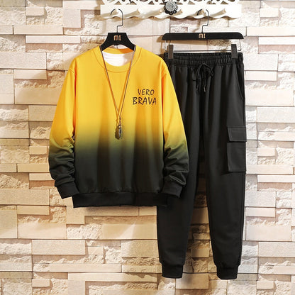 Completo sportivo uomo, felpa gialla sfumata con scritta "Vero Brava" e pantaloni neri cargo, appesi su sfondo muro mattoni.