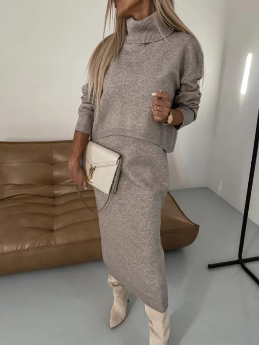 Donna con abito maglione grigio, borsa bianca e stivali beige, posa in un salotto moderno con divano marrone. Moda autunnale elegante.
