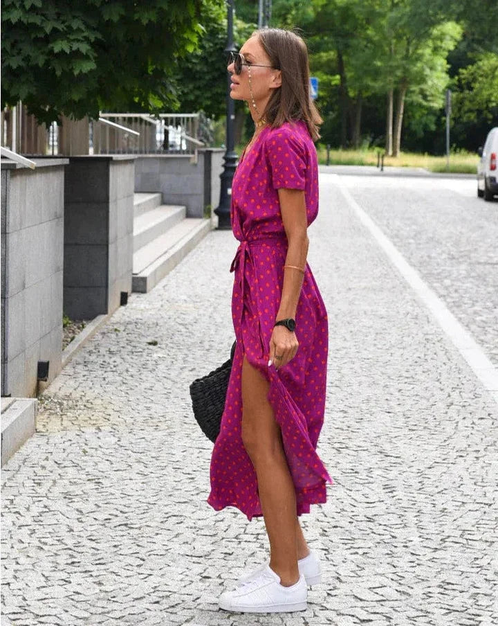 Donna in abito rosa a pois, occhiali da sole, borsa nera, scarpe da ginnastica bianche, cammina su strada lastricata, estate, moda casual.