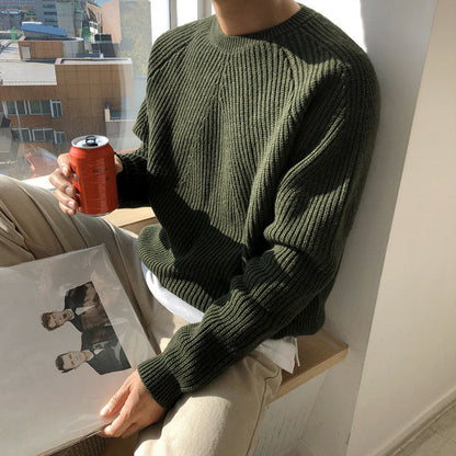 Uomo con maglione verde seduto vicino a finestra, tiene lattina di bibita e disco in vinile. Look casual, atmosfera rilassata, luce naturale.