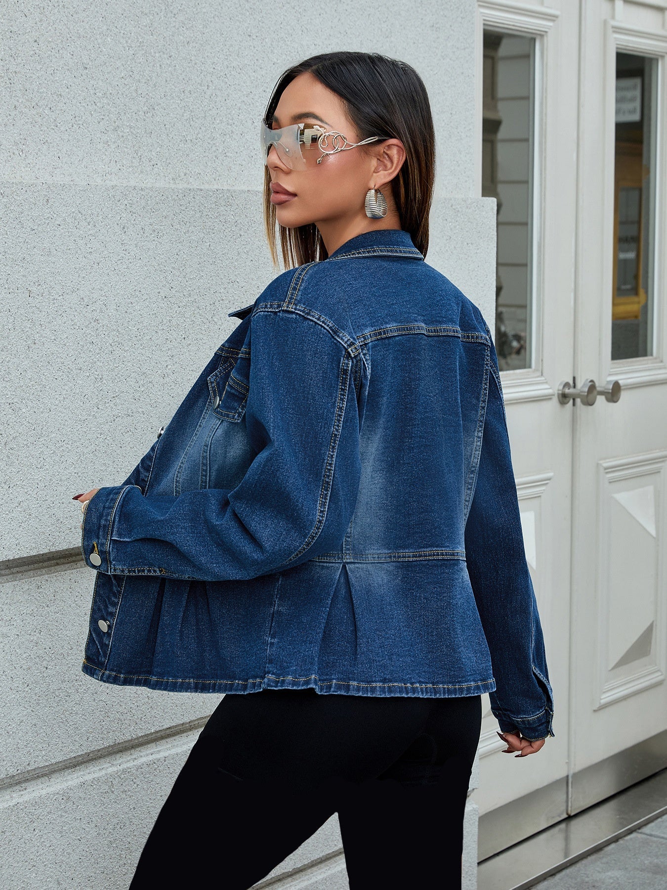 Donna con giacca di jeans blu, occhiali da sole specchiati e orecchini pendenti, posa all'aperto vicino a una porta bianca. Moda casual urbana.