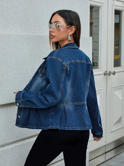 Donna con giacca di jeans blu, occhiali da sole specchiati e orecchini pendenti, posa all'aperto vicino a una porta bianca. Moda casual urbana.
