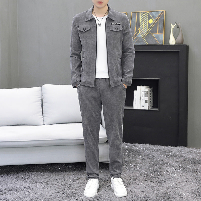 Uomo in tuta grigia elegante, in piedi su tappeto grigio, davanti a divano bianco e libreria moderna. Abbigliamento casual, moda maschile.