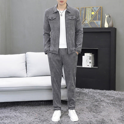 Uomo in tuta grigia elegante, in piedi su tappeto grigio, davanti a divano bianco e libreria moderna. Abbigliamento casual, moda maschile.