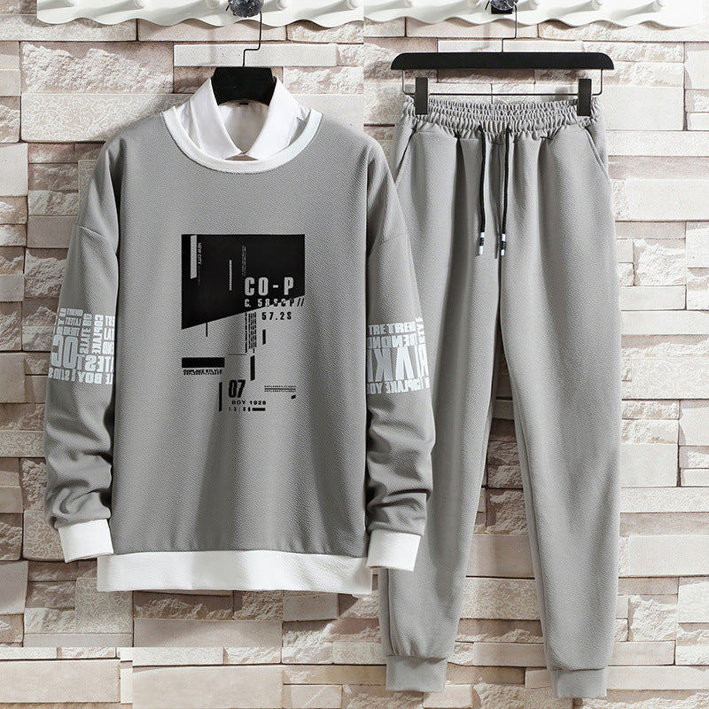 Completo tuta grigio da uomo con stampa grafica, felpa con colletto bianco e pantaloni con coulisse, appeso su sfondo di mattoni chiari.