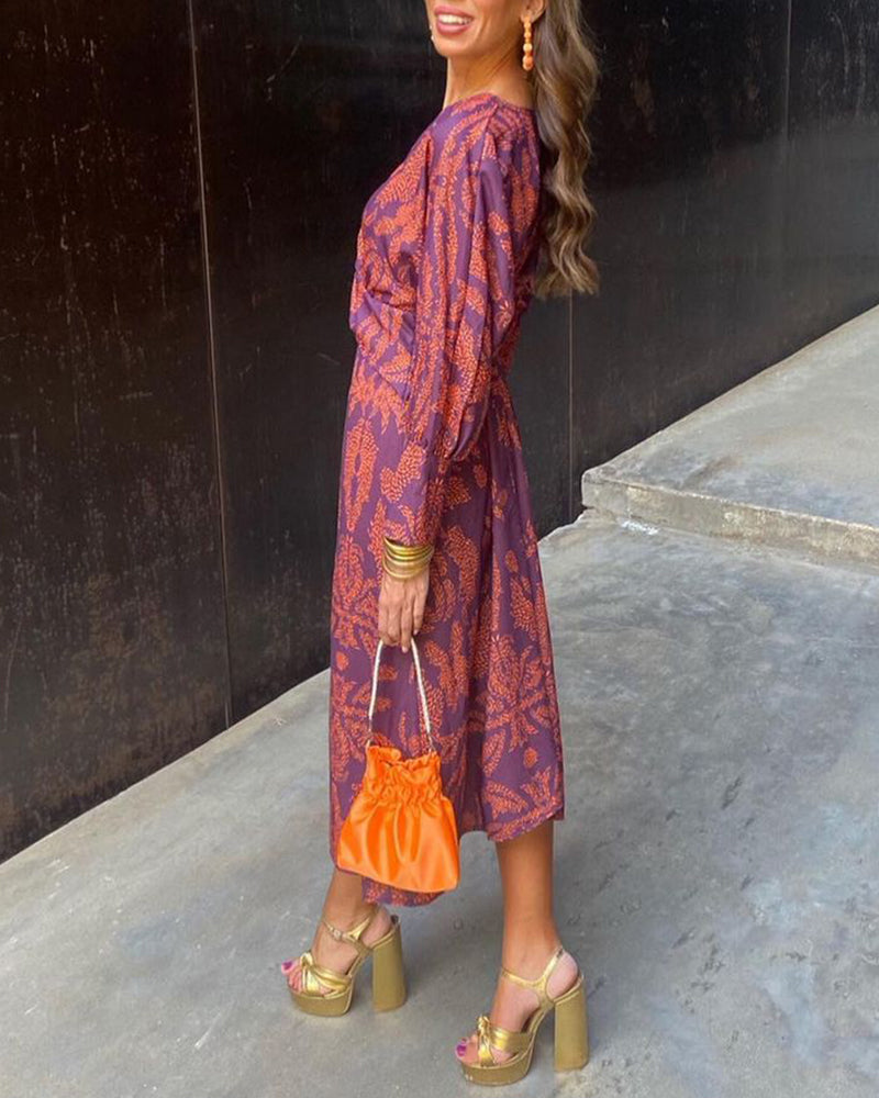 Donna in abito viola con motivi arancioni, borsa arancione e sandali dorati con tacco alto. Moda elegante, stile autunnale, accessori colorati.