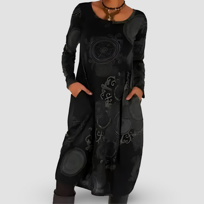 Abito lungo nero con motivi floreali, maniche lunghe, tasche laterali, indossato con collana. Elegante moda donna autunno-inverno.