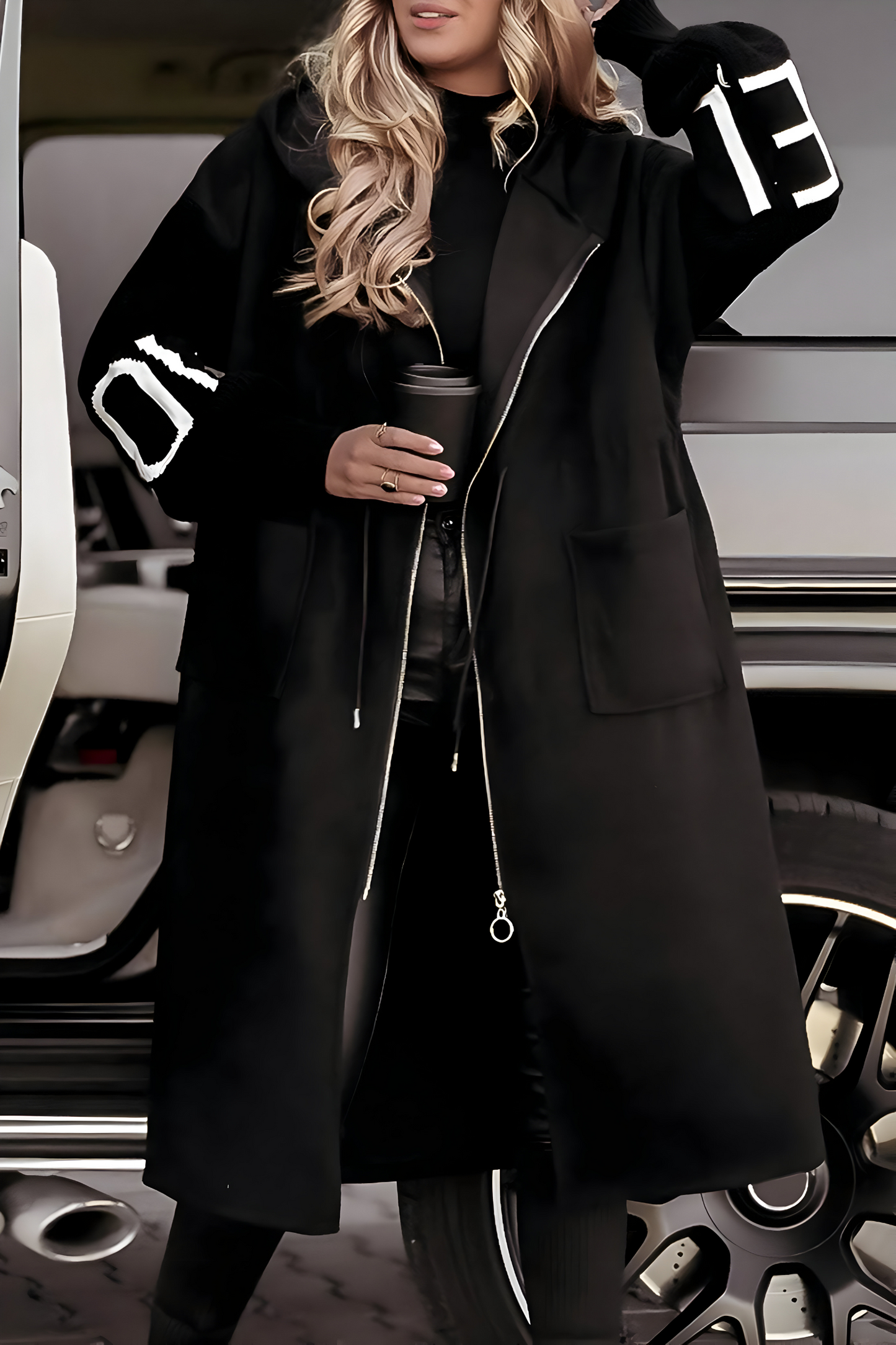 Donna con cappotto nero lungo, dettagli bianchi sulle maniche, tiene una tazza da caffè. Moda invernale, stile urbano, look casual chic.