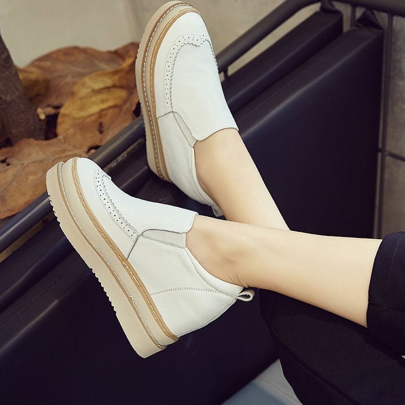 Scarpe slip-on bianche da donna, design casual con suola spessa, indossate su un divano nero. Scarpe moda comode, ideali per look quotidiani.