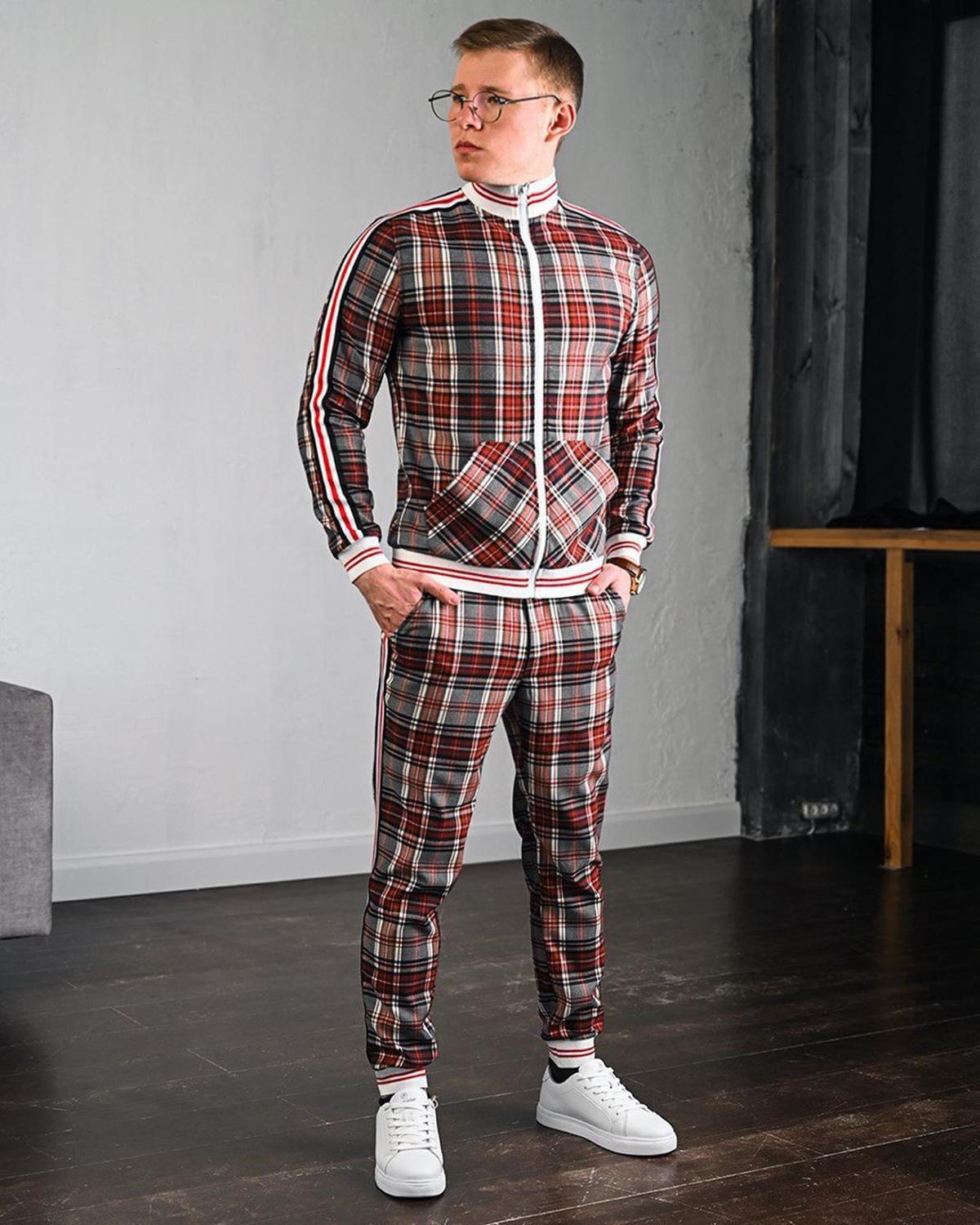 Giovane uomo in tuta a quadri rossi e bianchi, con occhiali e scarpe bianche, in posa su pavimento in legno. Moda maschile casual elegante.